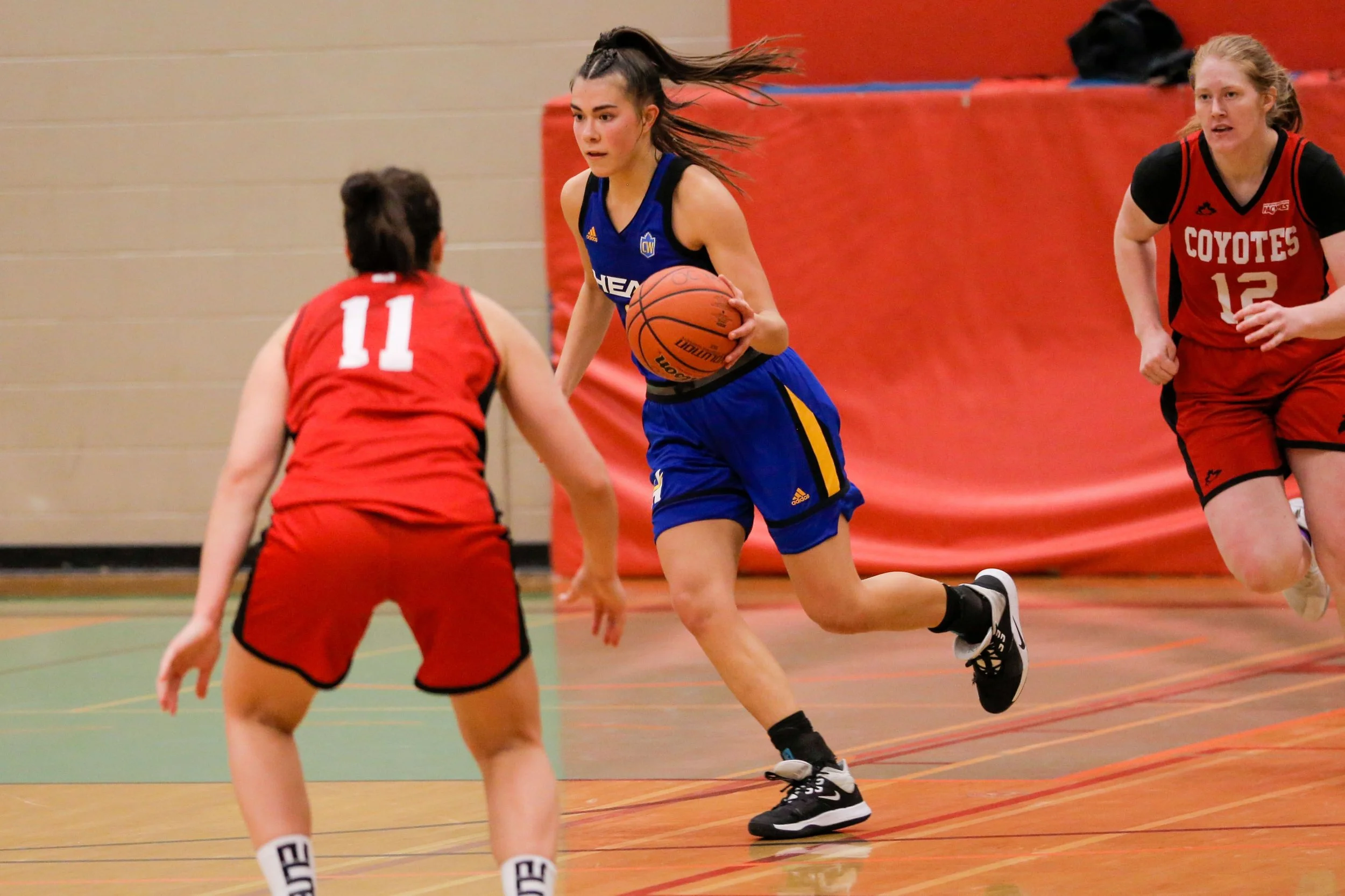 A girl in a blue basketball uniform dribbling a basketball while moving past two girls in red uniforms on an indoor basketball court.