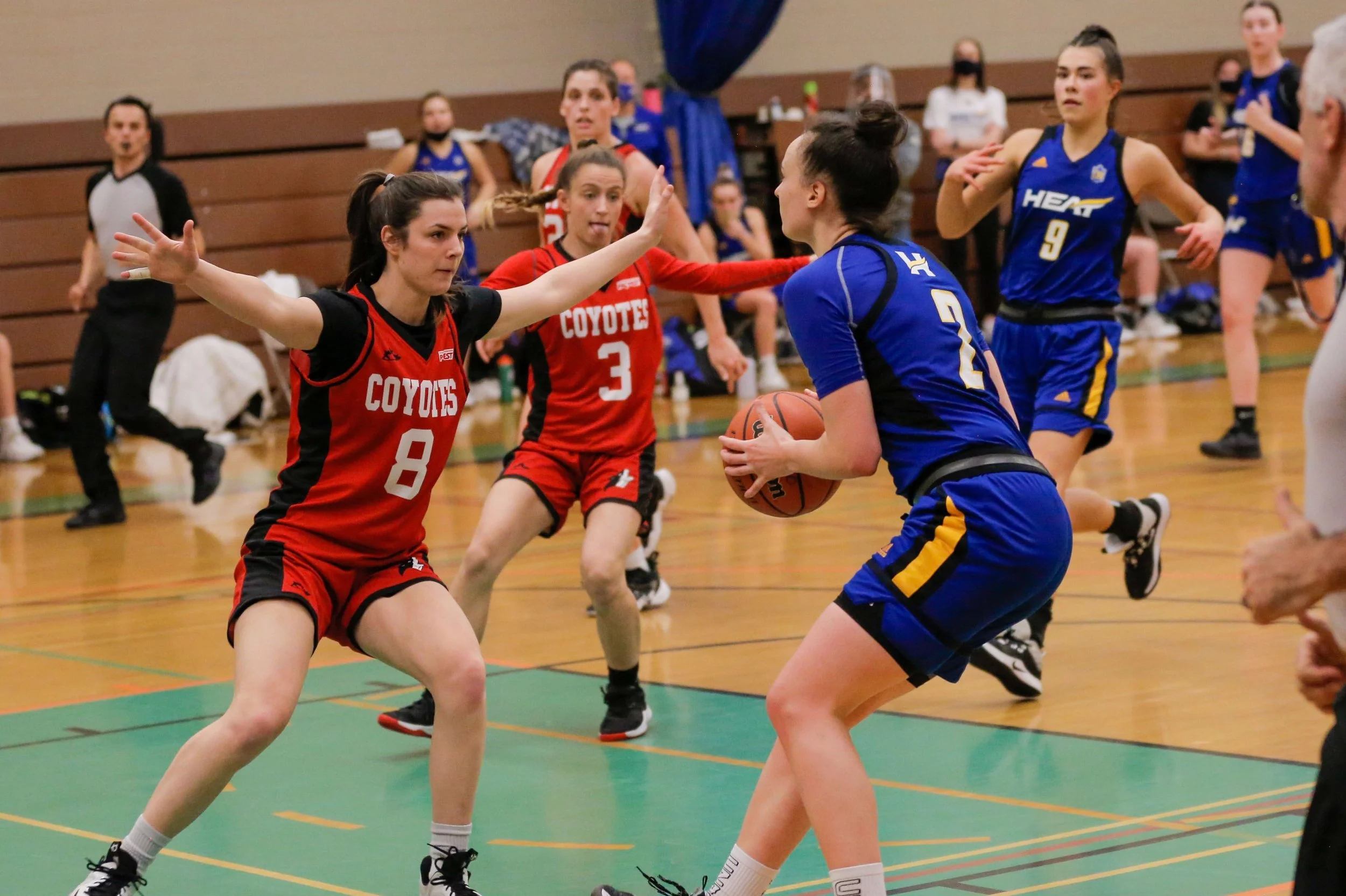 Young female basketball player in blue uniform with yellow accents holding a basketball, guarded by two opponents in red uniforms with black accents on a gymnasium court.