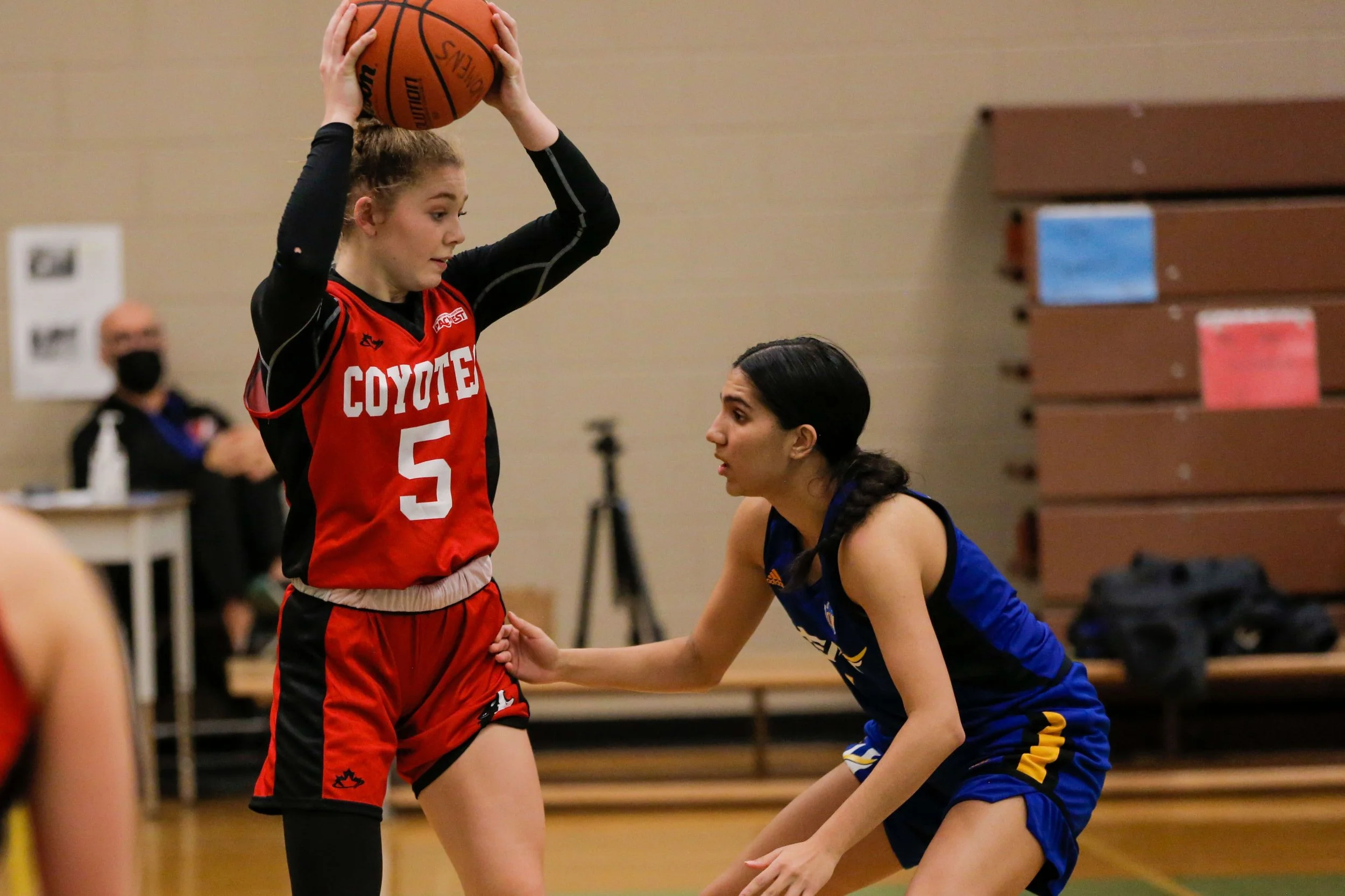 A female basketball player in a red uniform holding a basketball above her head while being guarded by another player in a blue uniform during a game.