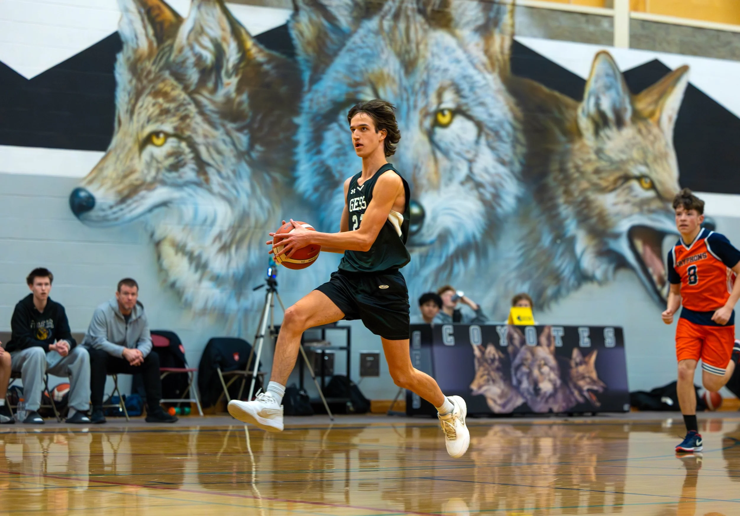 A young male basketball player in a black jersey jumps to shoot the ball during a game at a school gym. Behind him is a mural of three wolves on the wall. Other players and spectators are visible in the background.