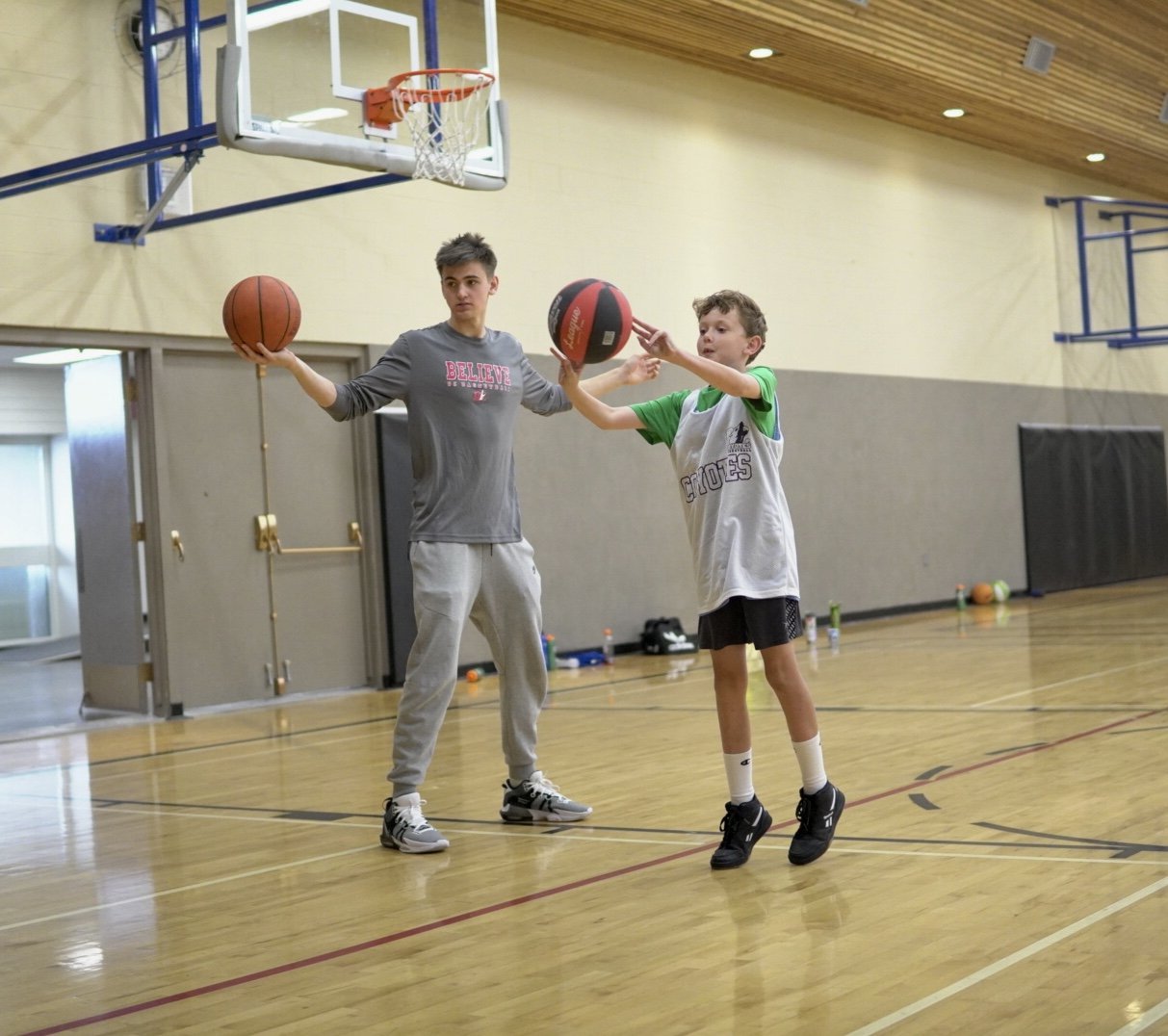 A young boy and a teenage coach playing basketball in an indoor gym. The coach holds a basketball while the boy reaches to catch a second ball. The gym has a wooden floor, a basketball hoop, and sports equipment scattered around.