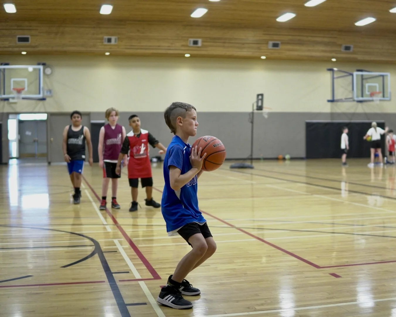 A young boy in a blue shirt and black shorts practicing basketball shooting in an indoor gym with three kids in the background waiting in line.