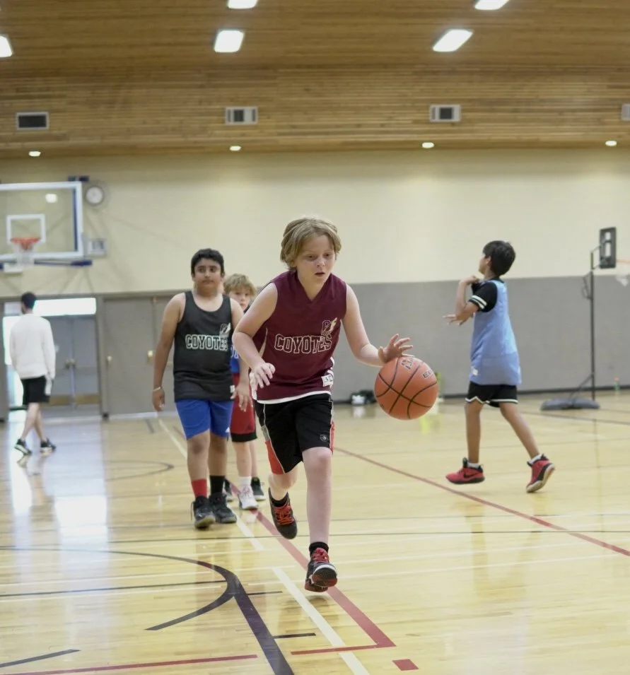 Children playing basketball in an indoor gym, with one child dribbling the basketball while others are in the background.