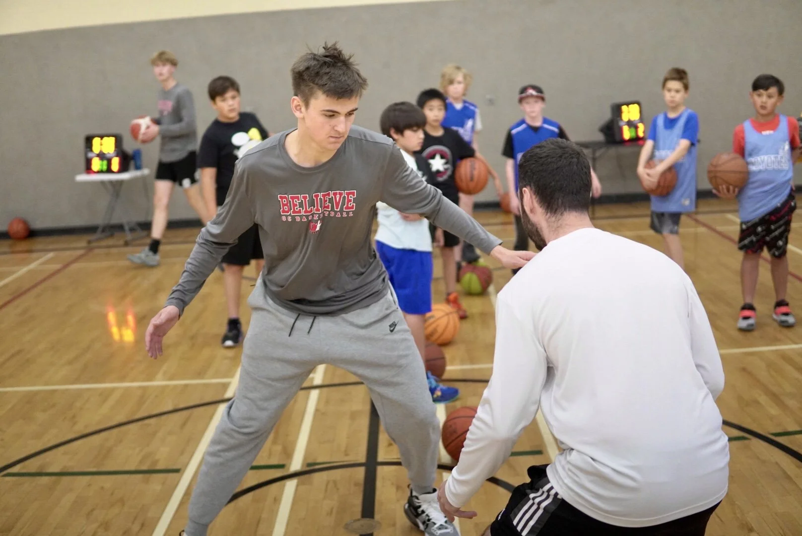 A basketball coach assisting a young player during a youth basketball practice session in a gym, with other children holding basketballs in the background.