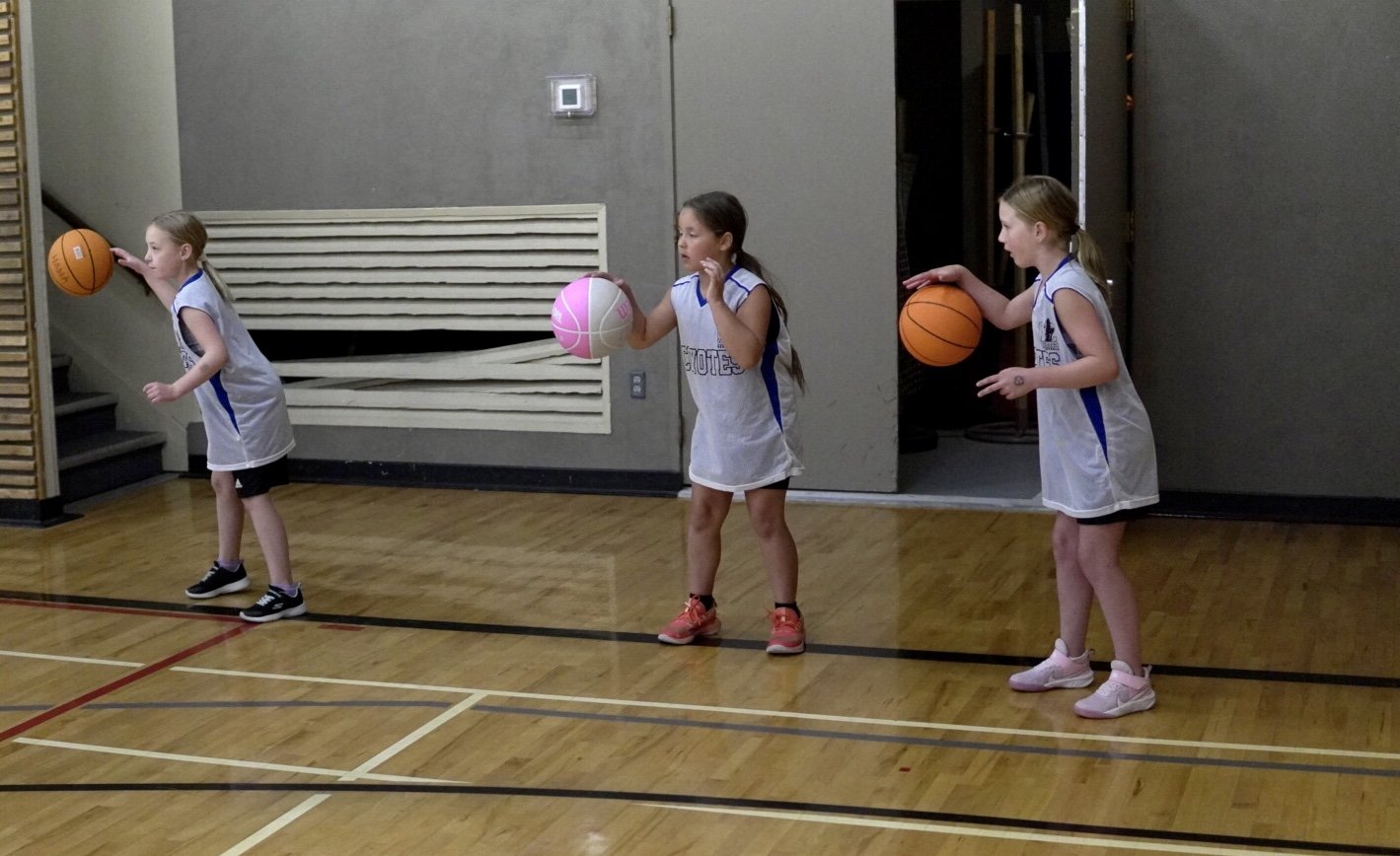 Three young girls in basketball uniforms practicing dribbling basketballs in a gymnasium with a hardwood floor.