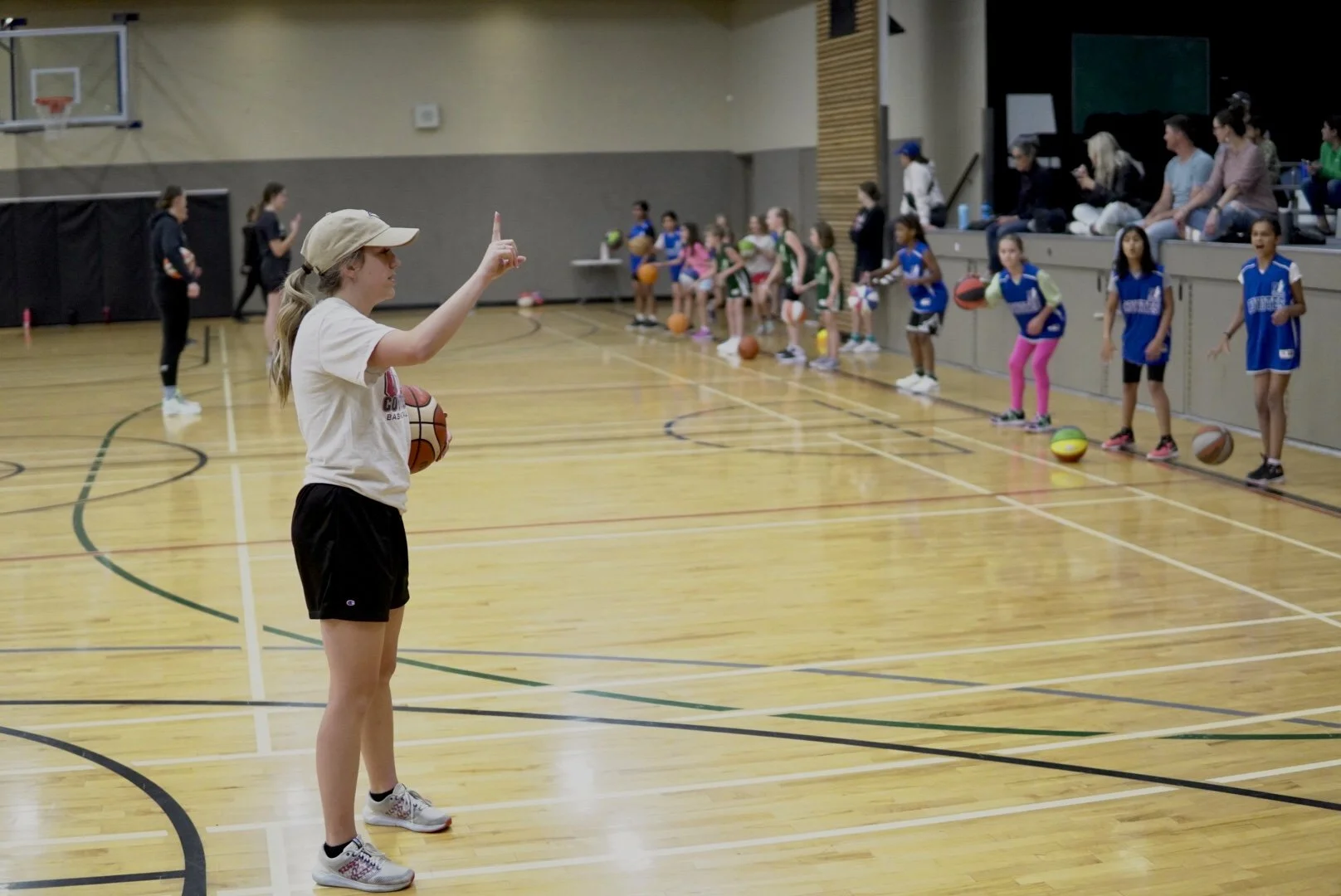 Girl in a beige hat and white T-shirt giving instructions in a gymnasium during a youth basketball practice, with children holding basketballs and sitting on a bench, some in blue uniforms.