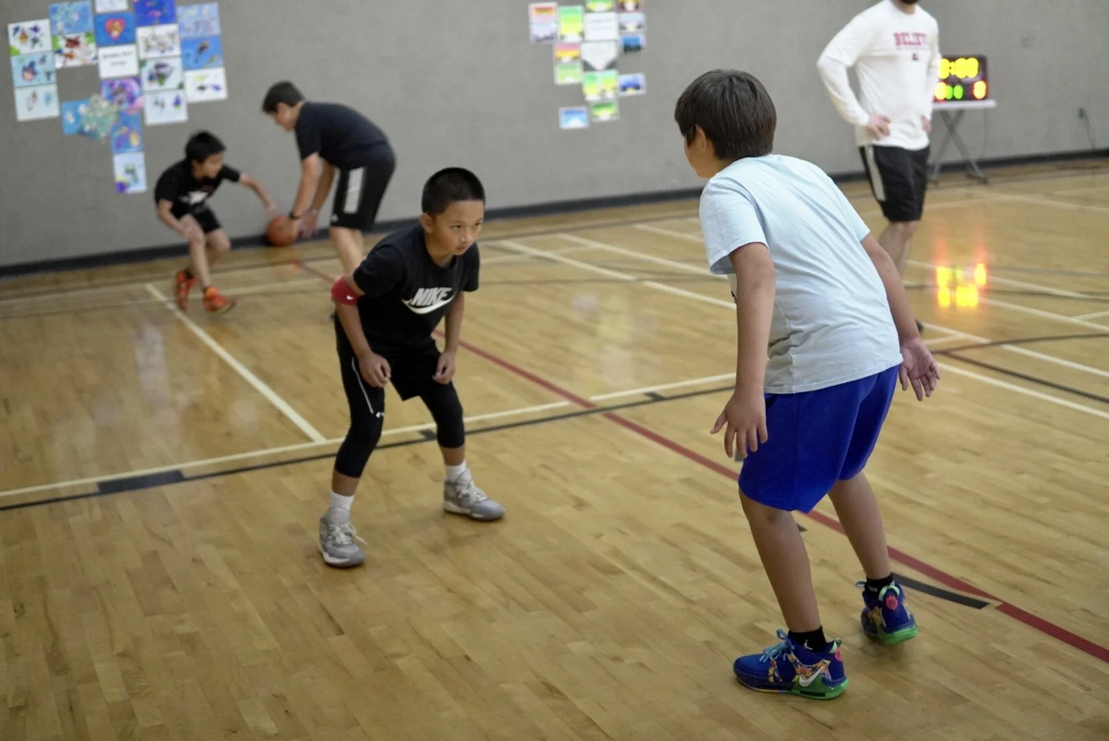 Children playing basketball in a gym, with two kids in the foreground competing and two in the background dribbling a basketball. An adult in a white shirt is also present.