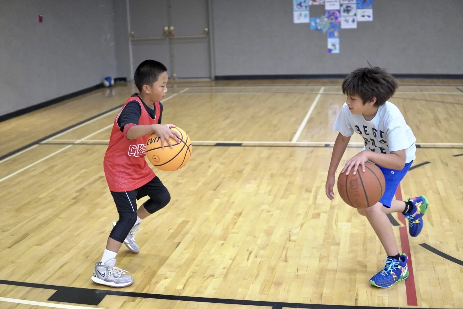 Two children playing basketball in a gym, one holding a yellow basketball and the other dribbling an orange basketball, on a wooden court.