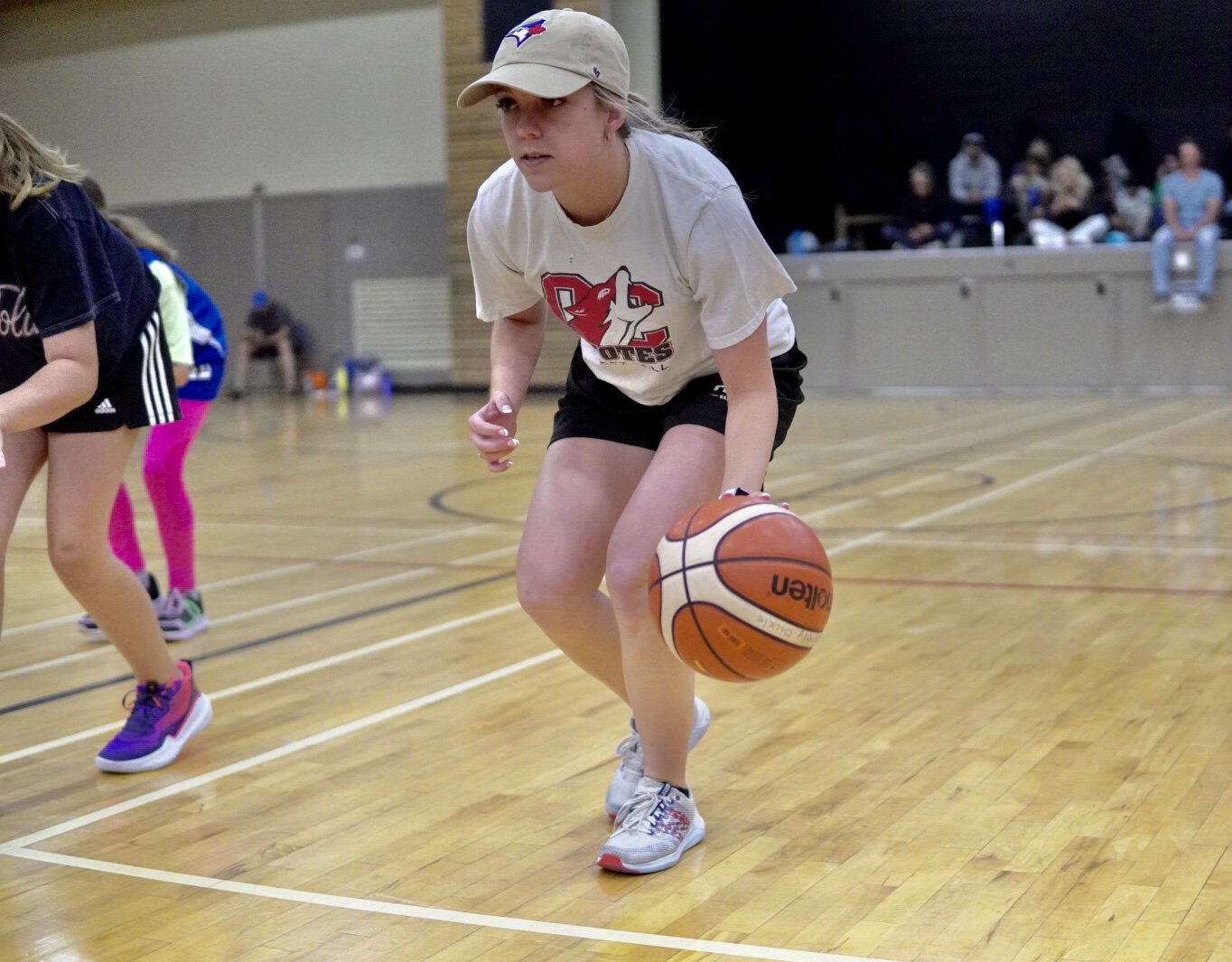 Young girl dribbling a basketball on an indoor court during a game or practice session, with other children and spectators in the background.