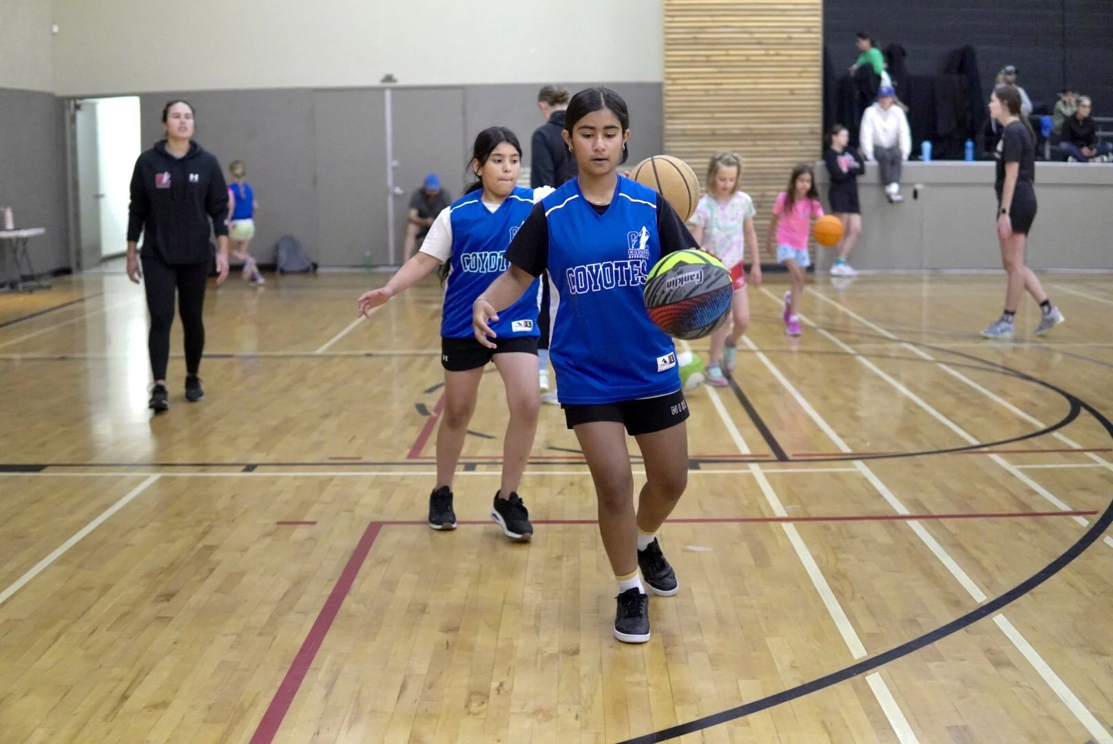 Young girl in a blue sports jersey practicing basketball indoors, with other children and adults in the background.