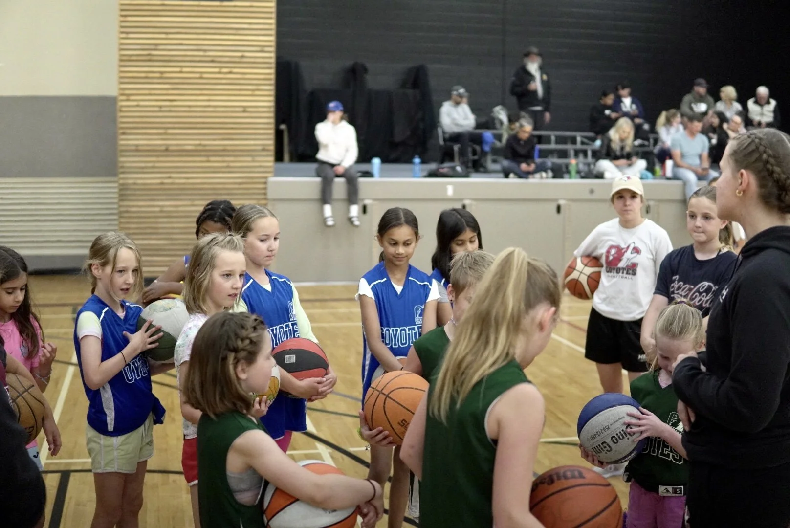 Group of young girls with basketballs listening to a female coach during a basketball practice in a gymnasium.