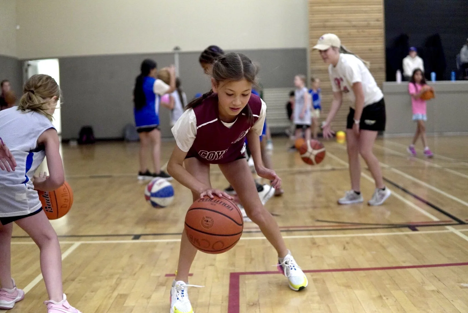 Young girls practicing basketball drills in an indoor gymnasium, dribbling basketballs.