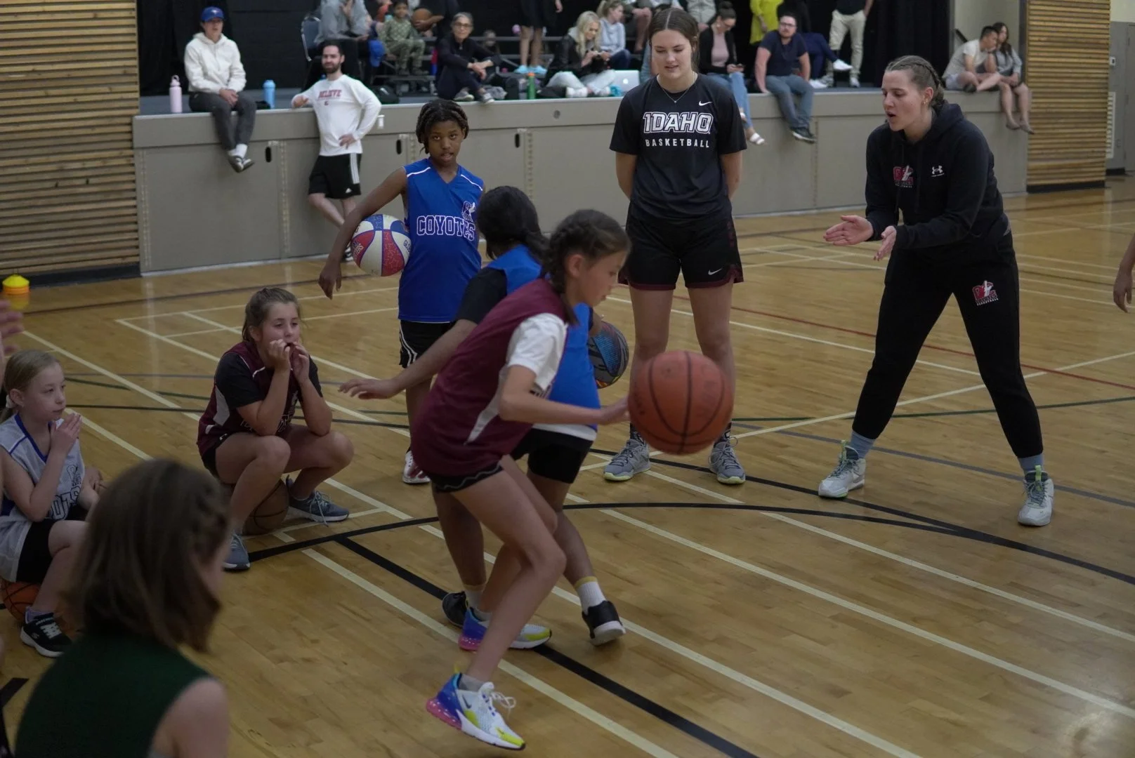 Young girls playing basketball on an indoor court while an adult coach instructs, with spectators seated in the background.