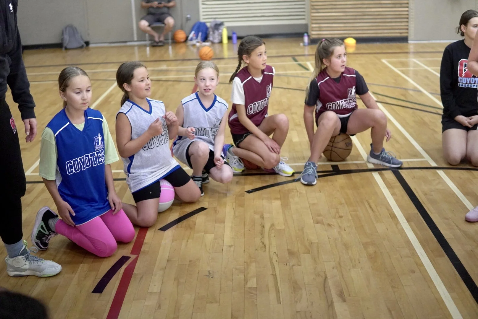 Young girls in basketball uniforms kneeling on a gym floor during a team meeting or ceremony.