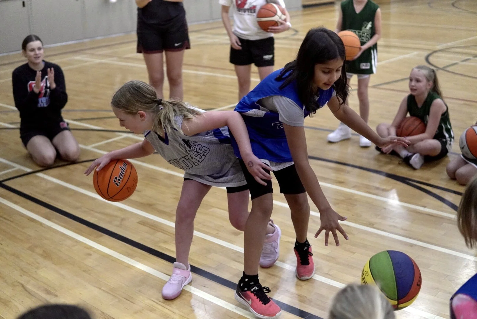 Young girls participating in a basketball training session in a gym, practicing dribbling with basketballs, supervised by an instructor.