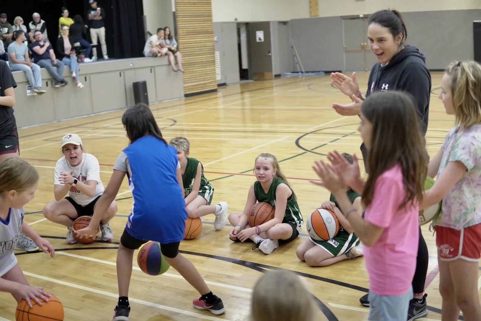 Kids participating in a basketball drill on a gym floor while a coach supervises and claps.