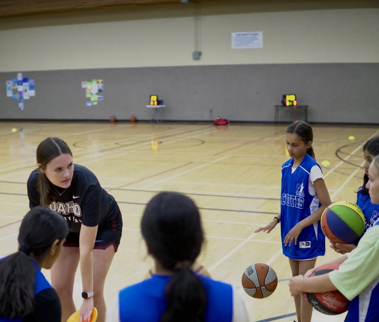 A basketball coach instructing young girls in basketball practice in a gymnasium, with some girls holding basketballs and listening to her, and others standing nearby.