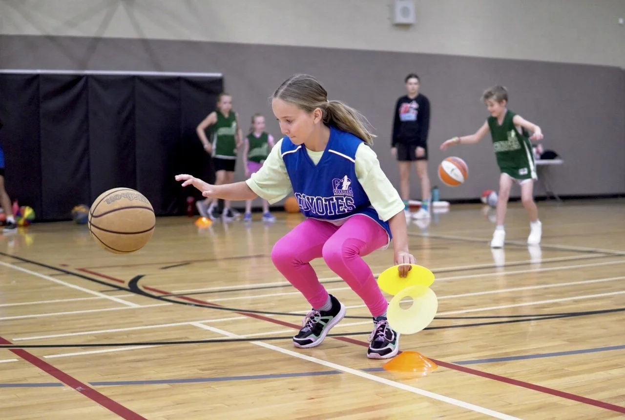Girl practicing basketball dribbling and agility drills in a gym, with other children in the background.