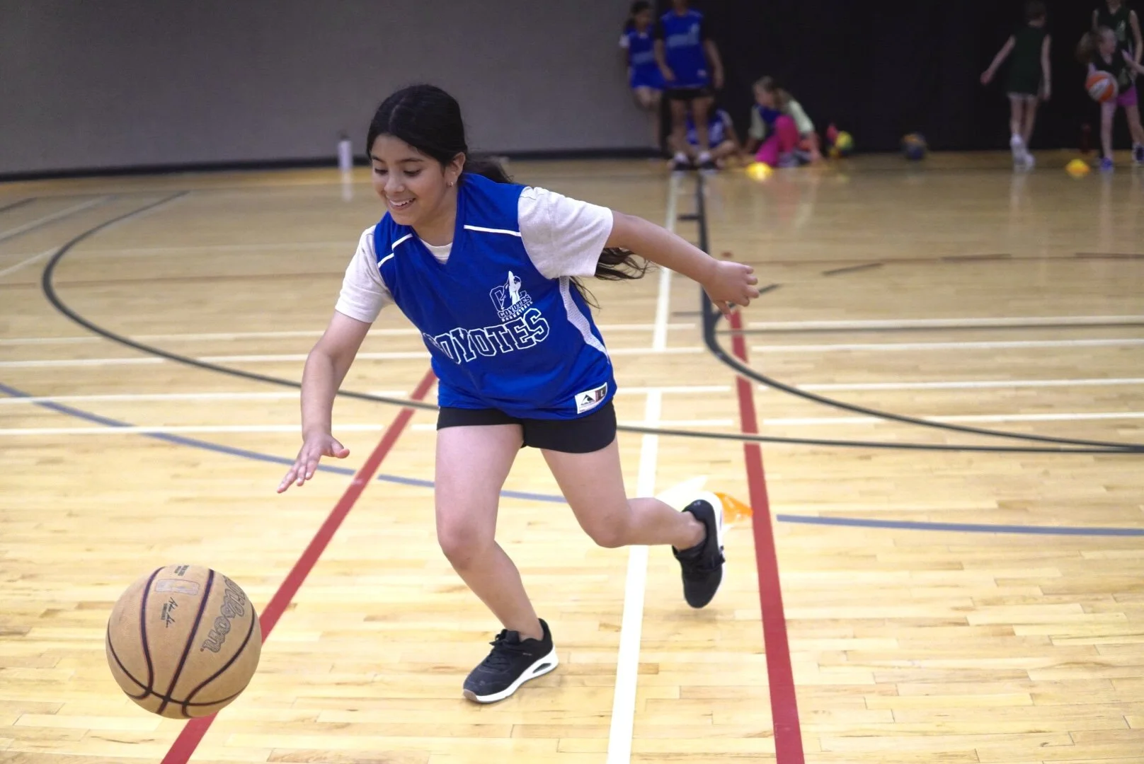 A young girl in a blue basketball jersey and black shorts dribbling a basketball on an indoor gym court.