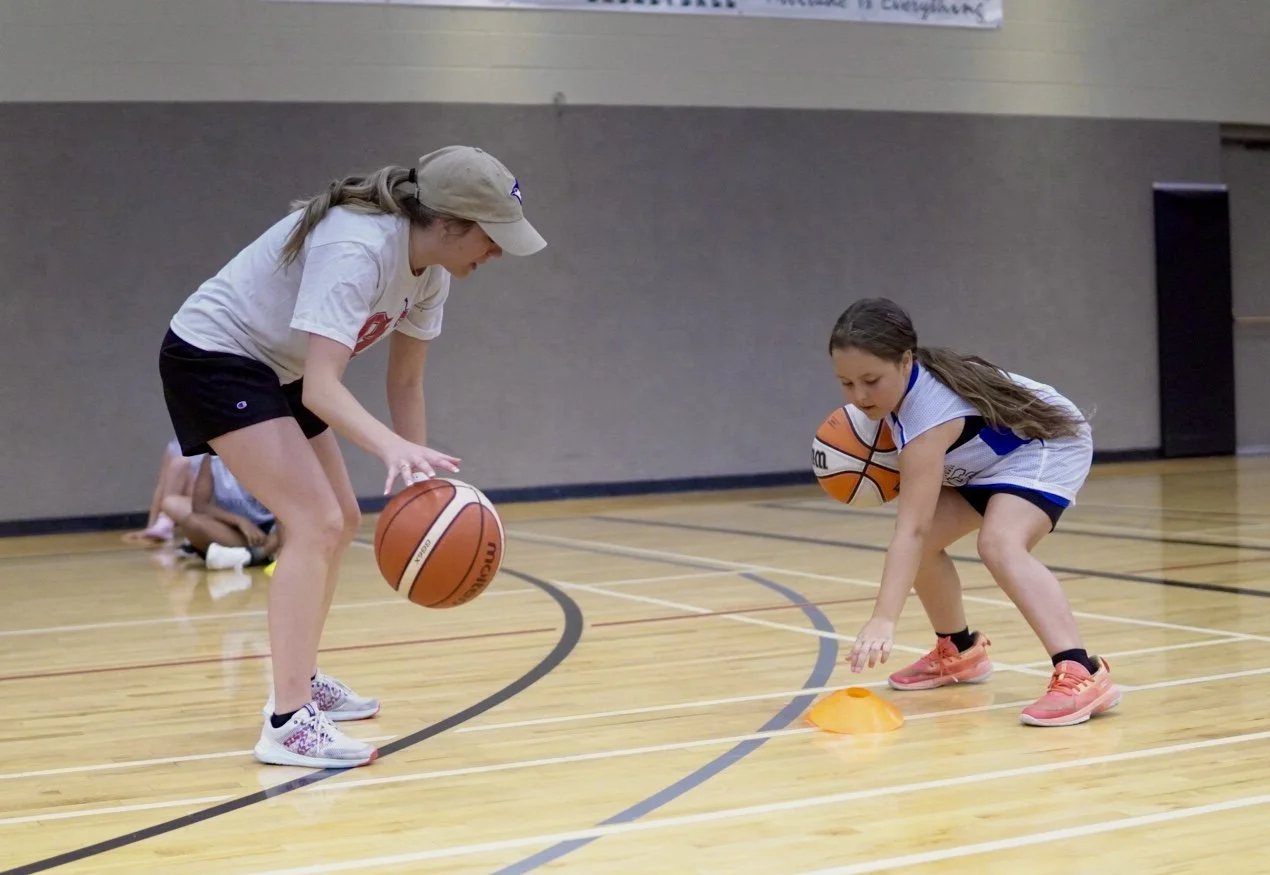 Two young girls practicing basketball dribbling exercises on an indoor court, with one girl dribbling a basketball and the other girl handling a basketball while crouching near an orange cone.
