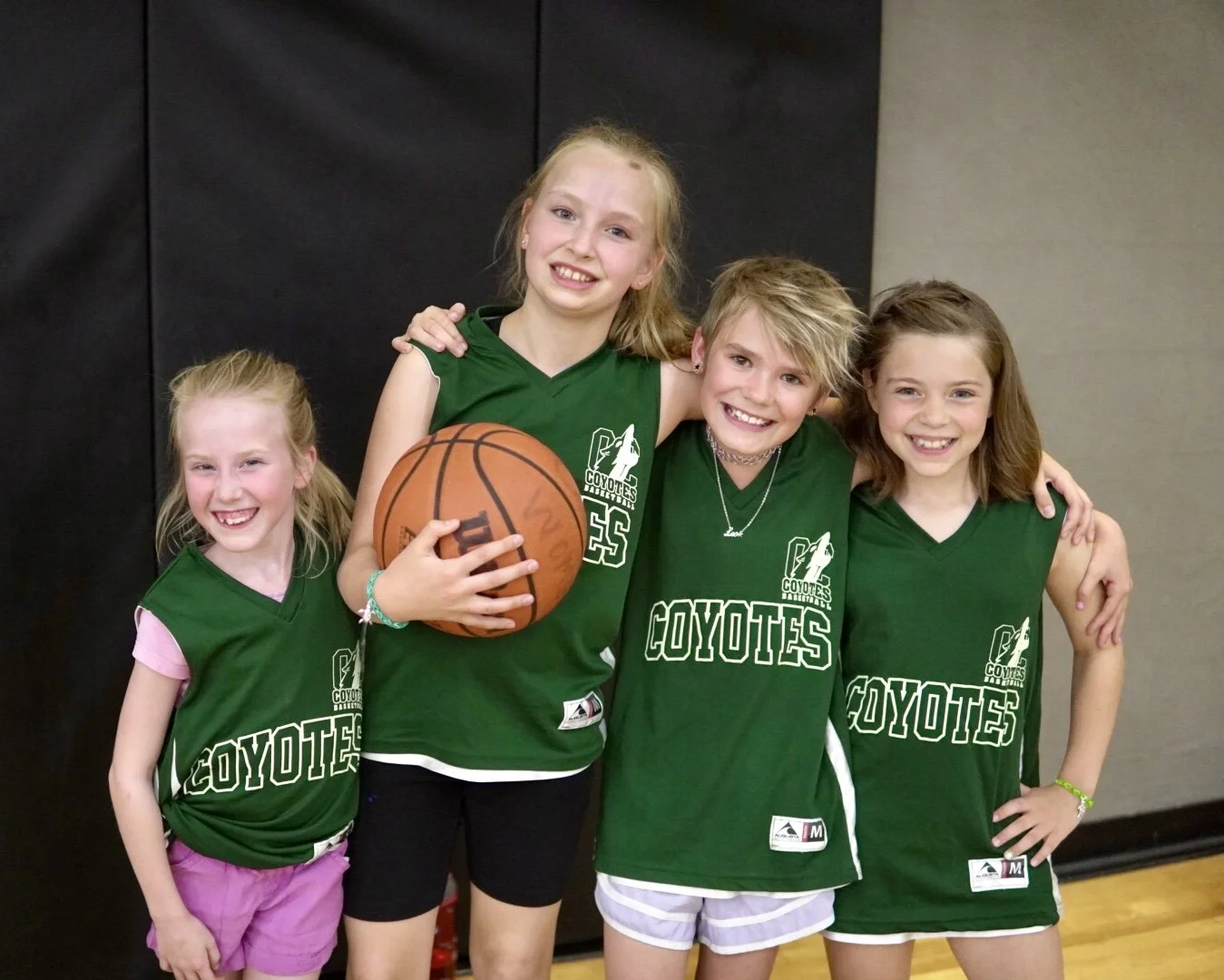 Four young girls wearing green basketball jerseys with 'Coyotes' written on them, smiling and posing together indoors, with one girl holding a basketball.
