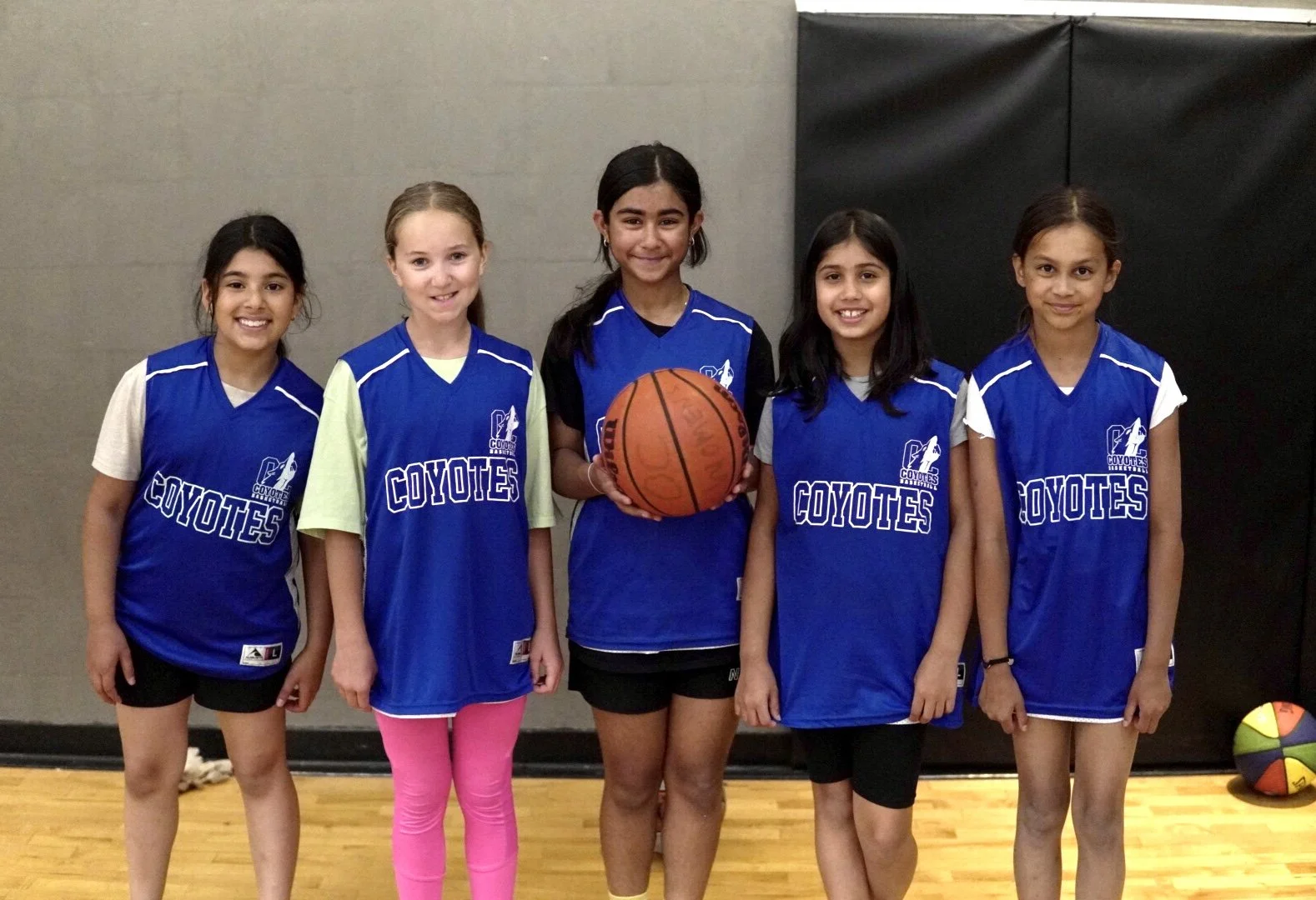 Group of five young girls in blue basketball jerseys posing in a gym, one holding a basketball.