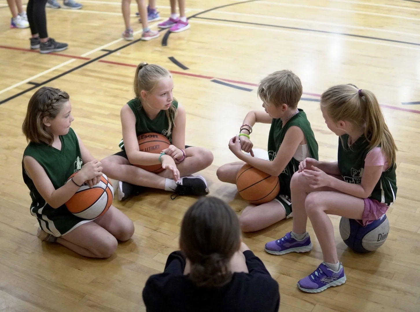 Four young girls in green basketball jerseys sitting on the gym floor, two holding basketballs, engaged in a discussion, with a coach or adult sitting in front of them.