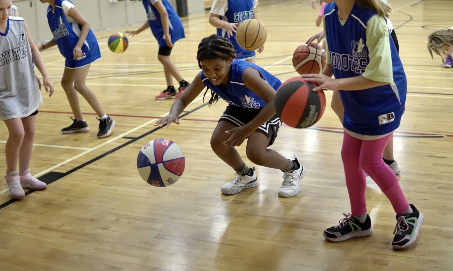 Children playing with basketballs and volleyballs on a gymnasium floor.