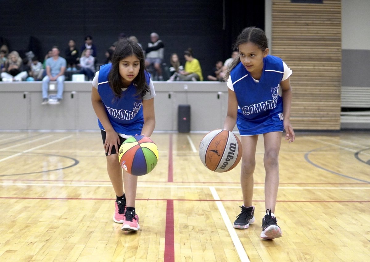 Two young girls in blue basketball jerseys practicing dribbling on an indoor basketball court, with spectators sitting in the background.