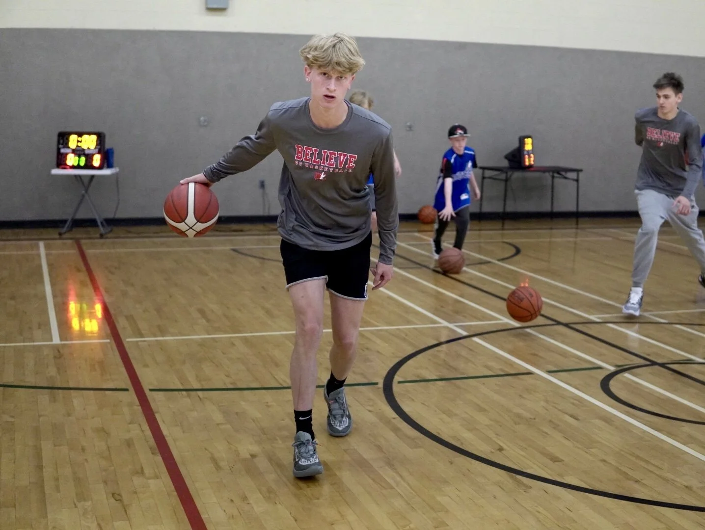A young man dribbling a basketball on an indoor court during a practice session with other children in the background.