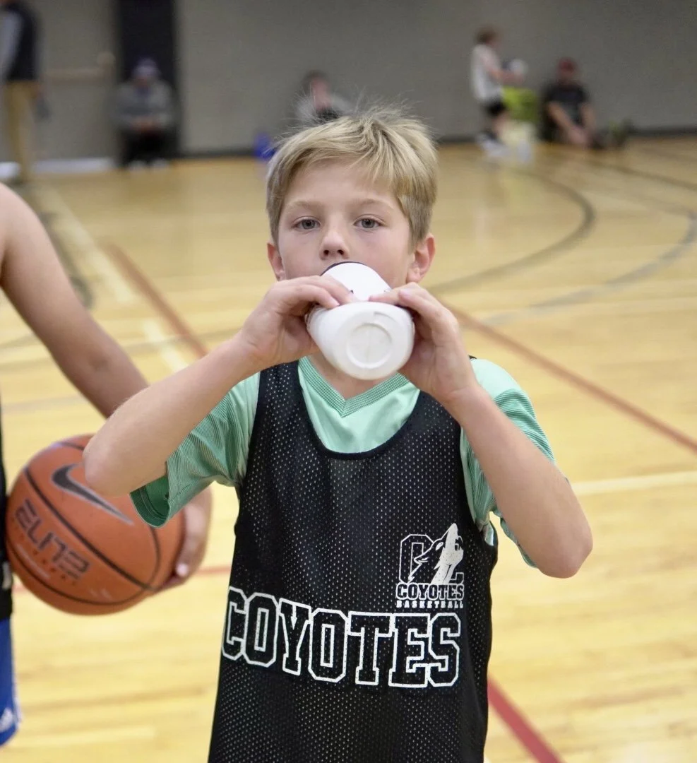 Young boy in a basketball jersey drinking from a white water bottle in a gymnasium, with other children and a basketball in the background.