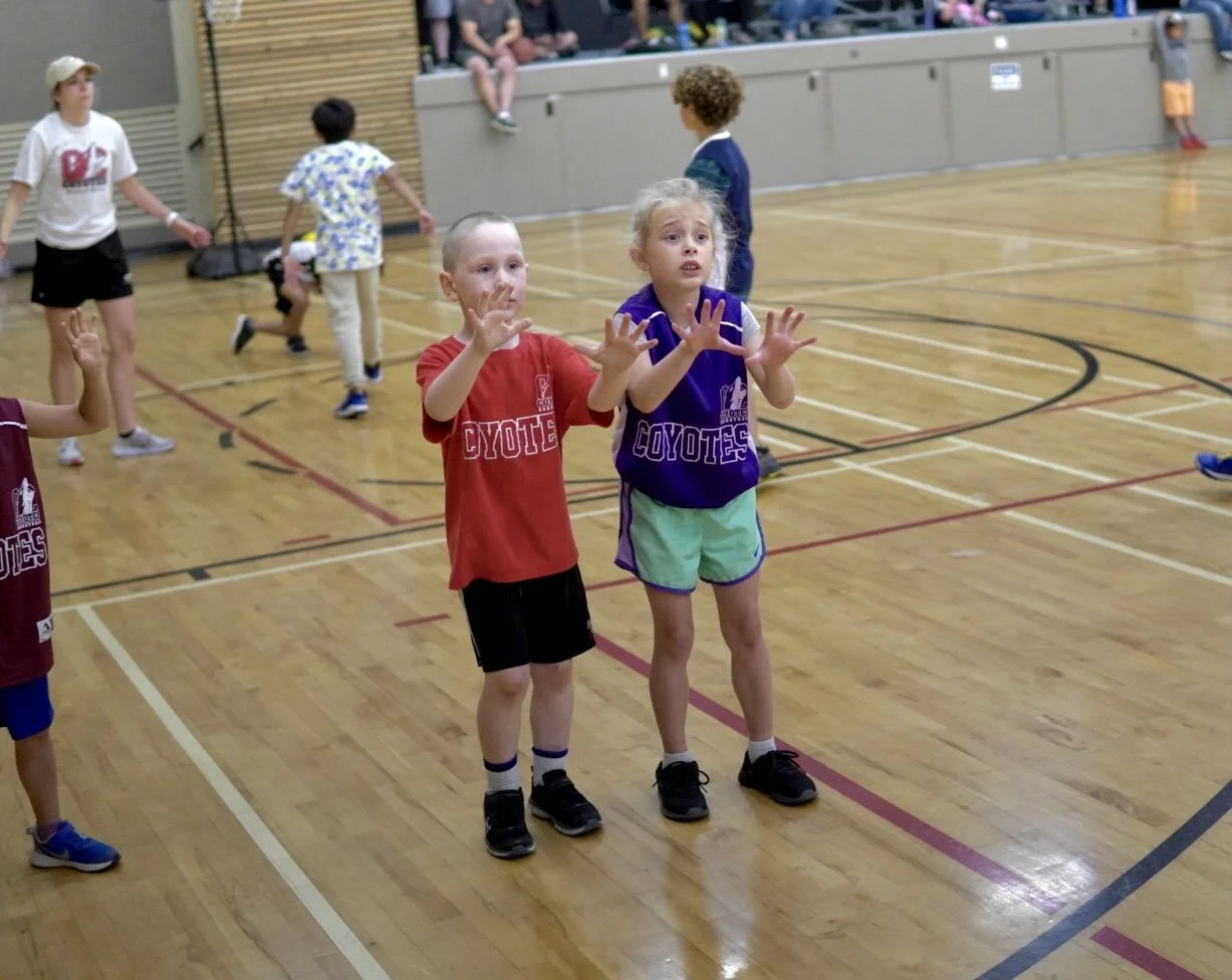 Children in a gymnasium playing a game, with two girls in the foreground raising their hands and looking distressed, wearing sports jerseys that say 'Coyote' in red and purple.