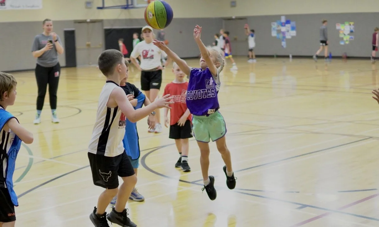 Children playing with a ball at an indoor gym, some wearing sports jerseys, with adults supervising in the background.