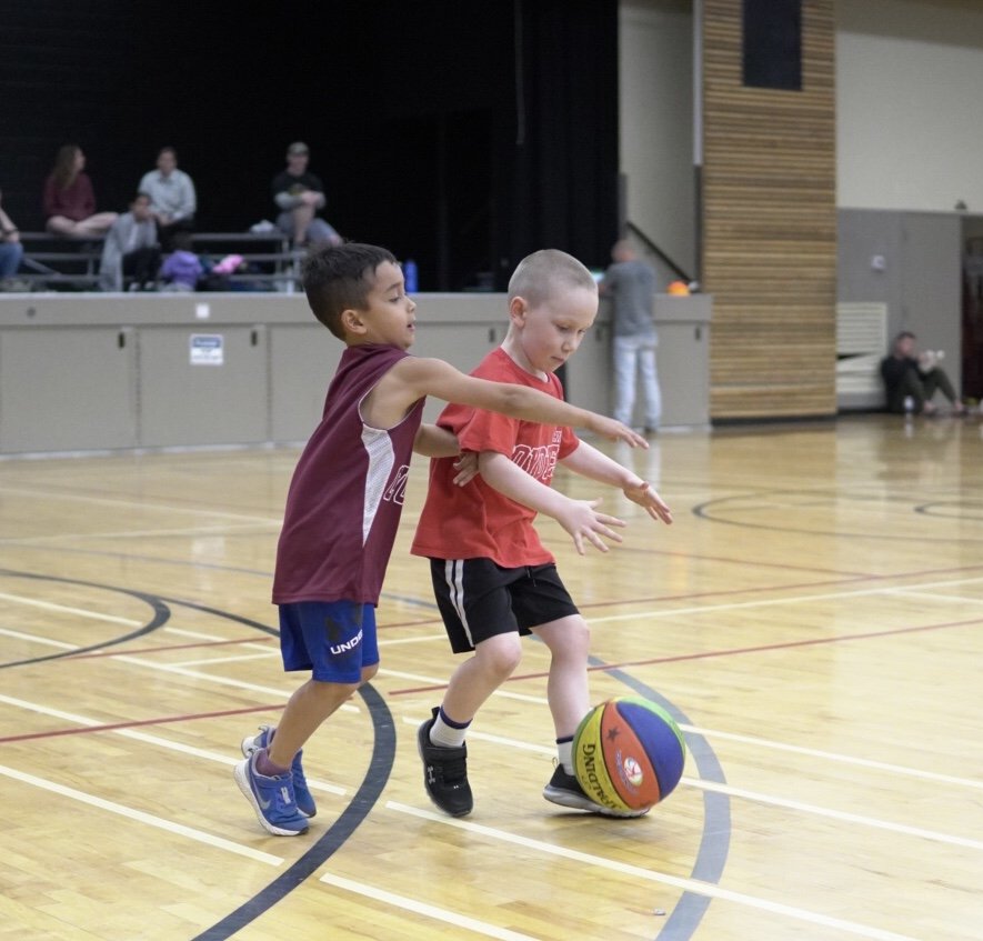 Two young boys playing basketball on an indoor court, with one boy in a maroon tank top and blue shorts trying to block the other boy in a red t-shirt and black shorts who is dribbling a colorful basketball.