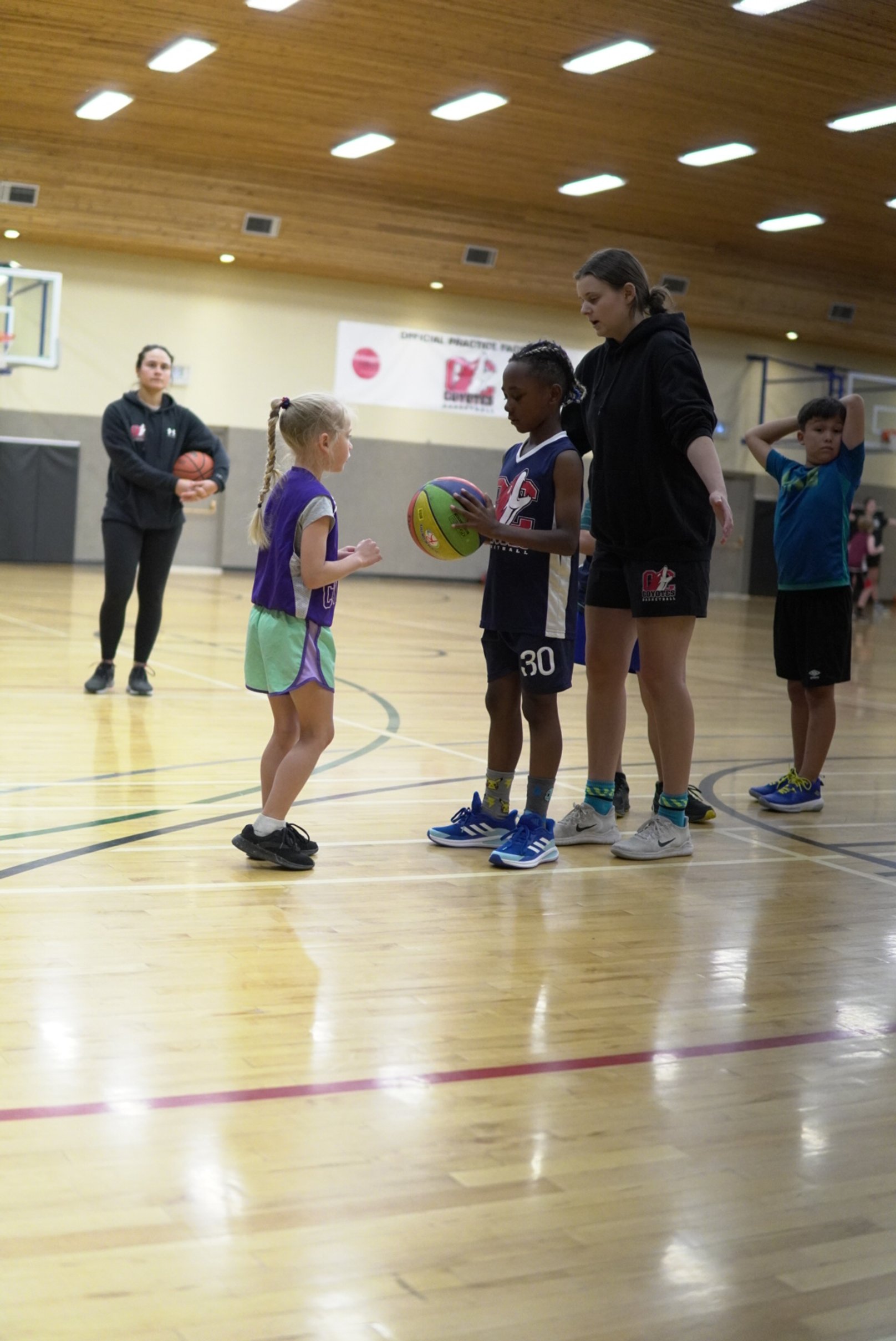 Young girl and woman on a basketball court with two boys in sportswear, one holding a basketball, in a gym with wooden flooring and ceiling, and a basketball hoop in the background.
