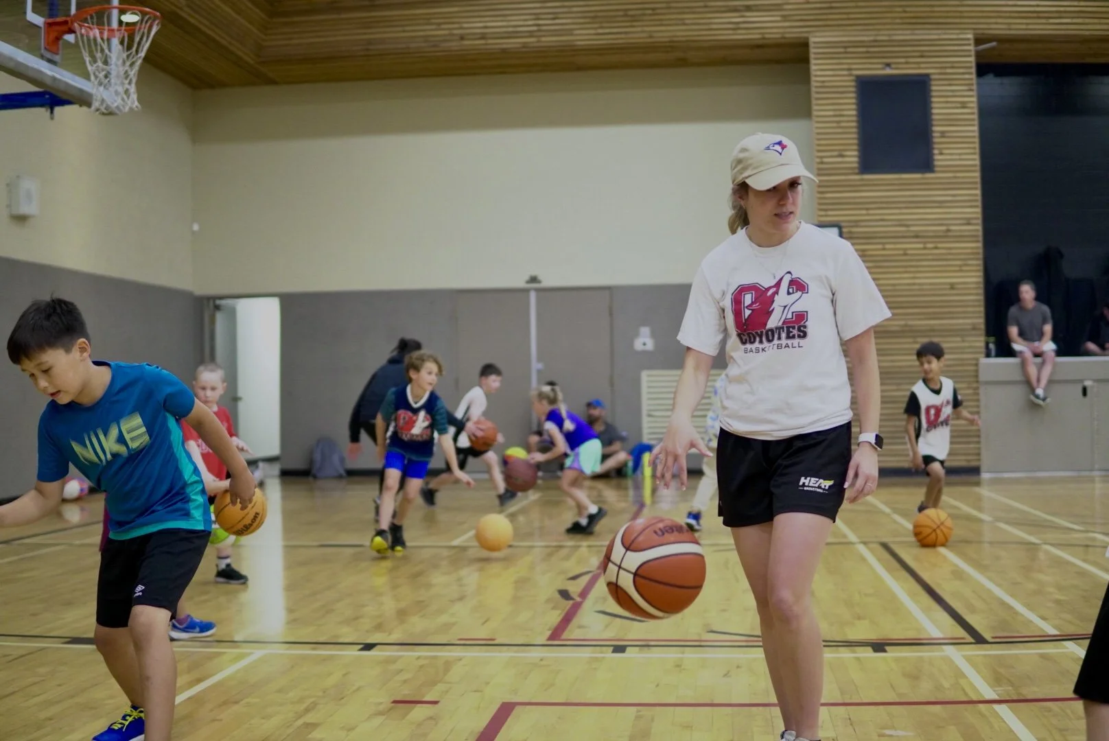 Children practicing basketball dribbling during a session in an indoor gym with a coach or instructor supervising.