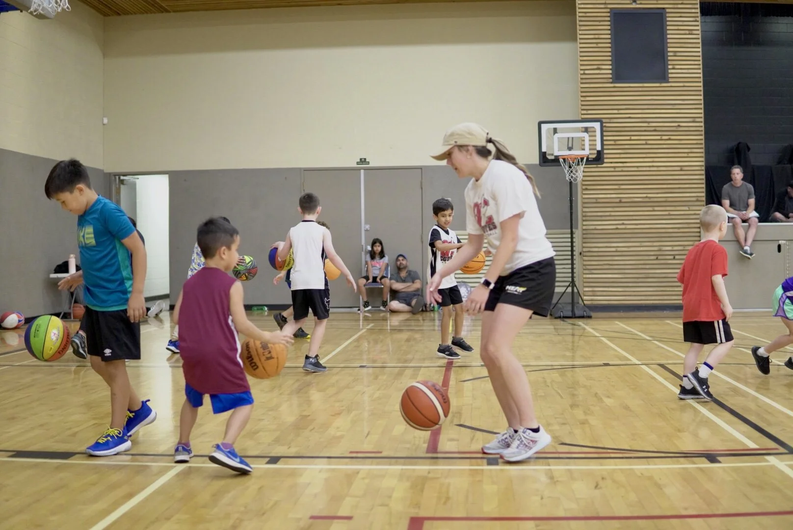 Children practicing basketball drills in an indoor gymnasium led by a female coach.