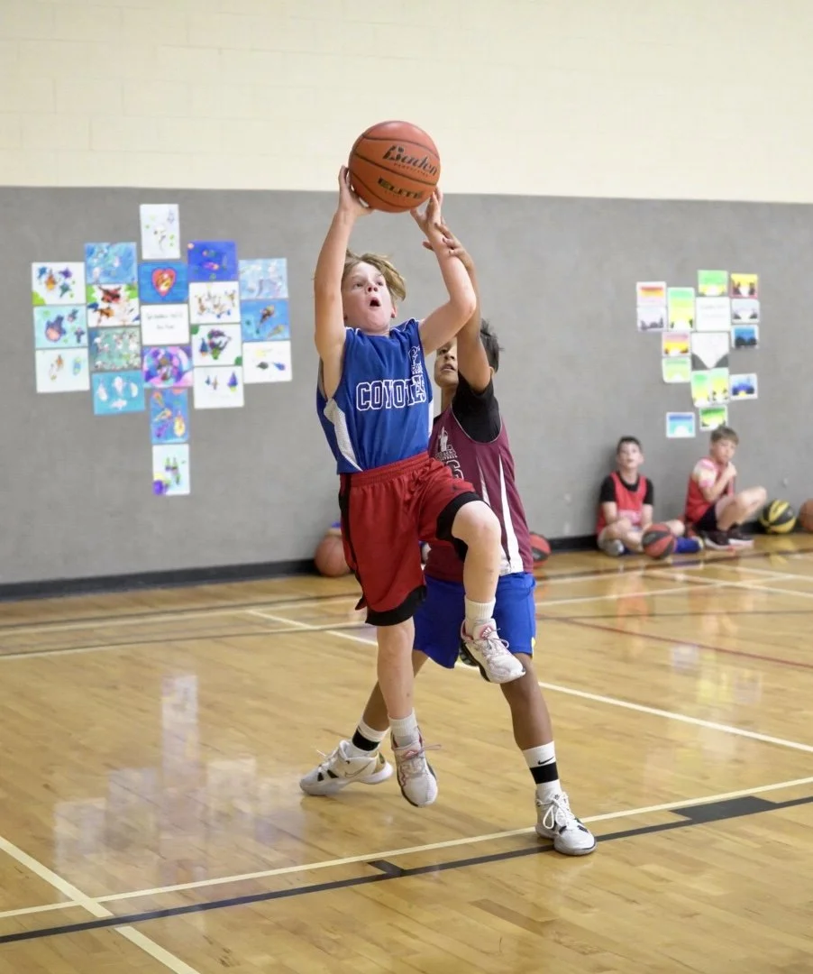 A young boy in a blue basketball jersey and red shorts is jumping to shoot a basketball in an indoor gym, with another child in a maroon jersey defending him. Other kids are seated on the floor in the background.