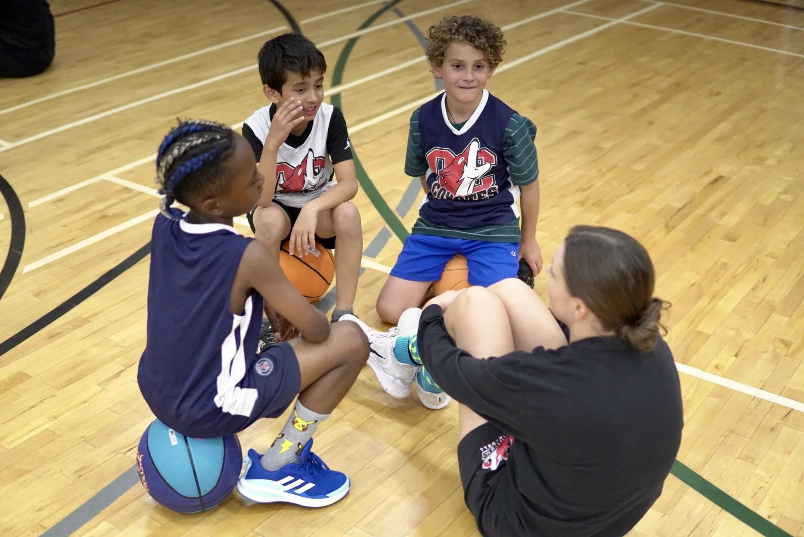 A group of young children on a basketball court, sitting in a circle, engaged in a discussion with a coach or adult. The children are wearing sports uniforms and are holding or sitting near basketballs.