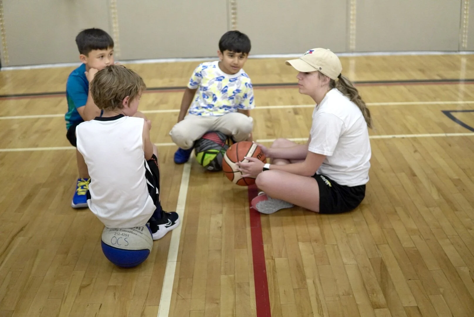 A female basketball coach sitting on the gym floor, holding a basketball, talking to four young boys who are sitting around her on the hardwood court.