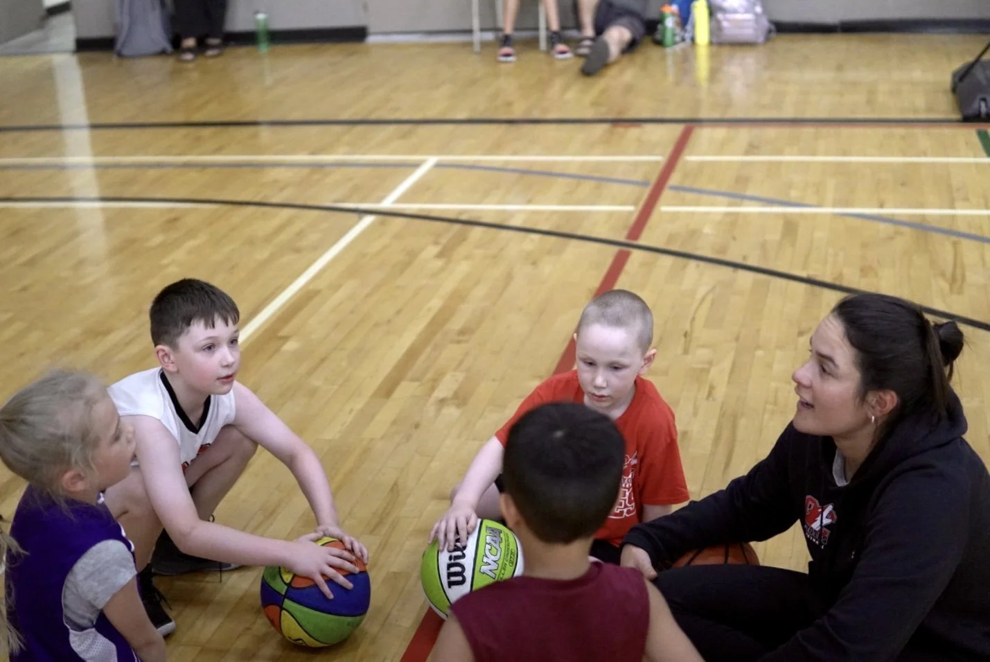 A group of young children and a coach sitting on a basketball court, engaged in a basketball practice or discussion.