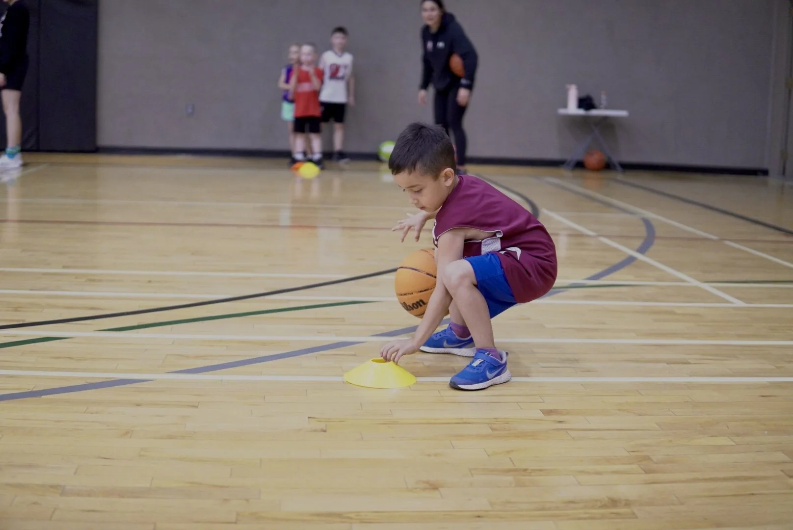 A young boy is squatting on a basketball court while practicing dribbling around a yellow cone, with other children and an adult in the background.