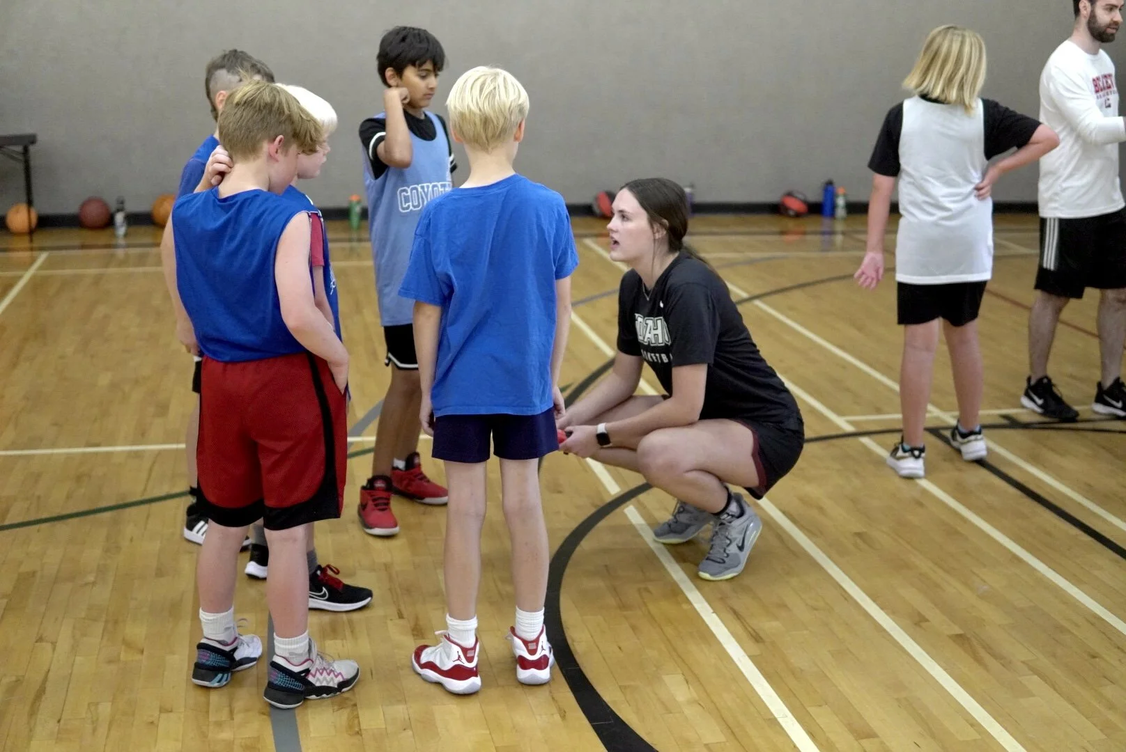 A female coach crouching and speaking to a group of young boys on a basketball court. The boys are standing in a semi-circle around her, listening attentively. Other children and a woman are visible in the background.