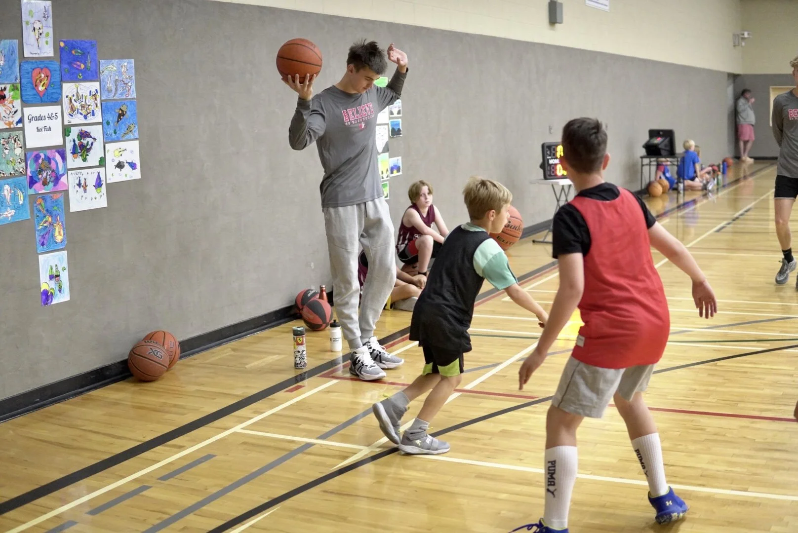 Young boys playing basketball in an indoor gym, with one boy holding a basketball above his shoulder. Two other boys are guarding him, and in the background, a girl sits on the floor near basketballs and sports bottles. Wall decorated with children's
