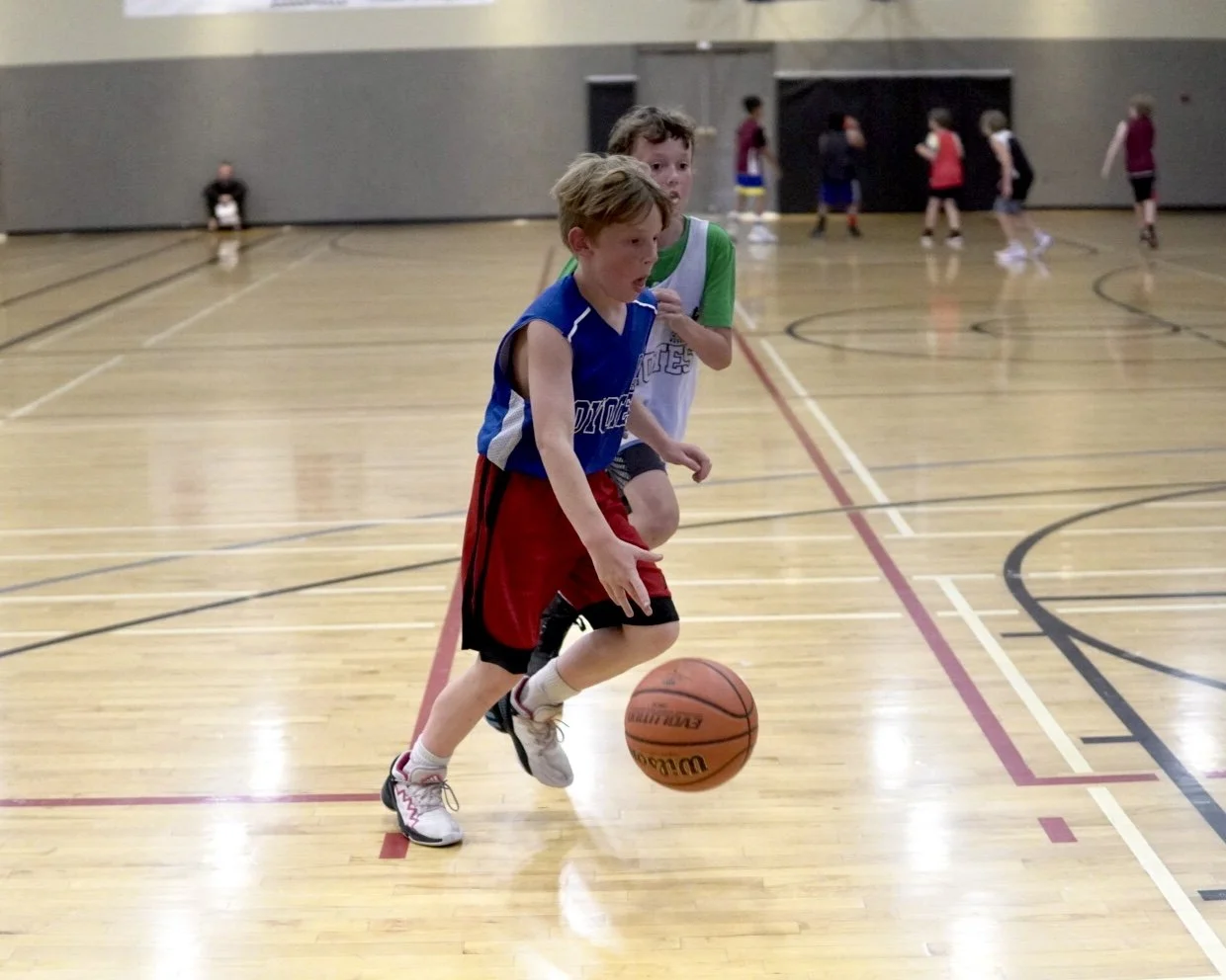 Two young boys playing basketball on an indoor court, one in a blue jersey and red shorts dribbling the ball, the other in a green and white jersey behind him.