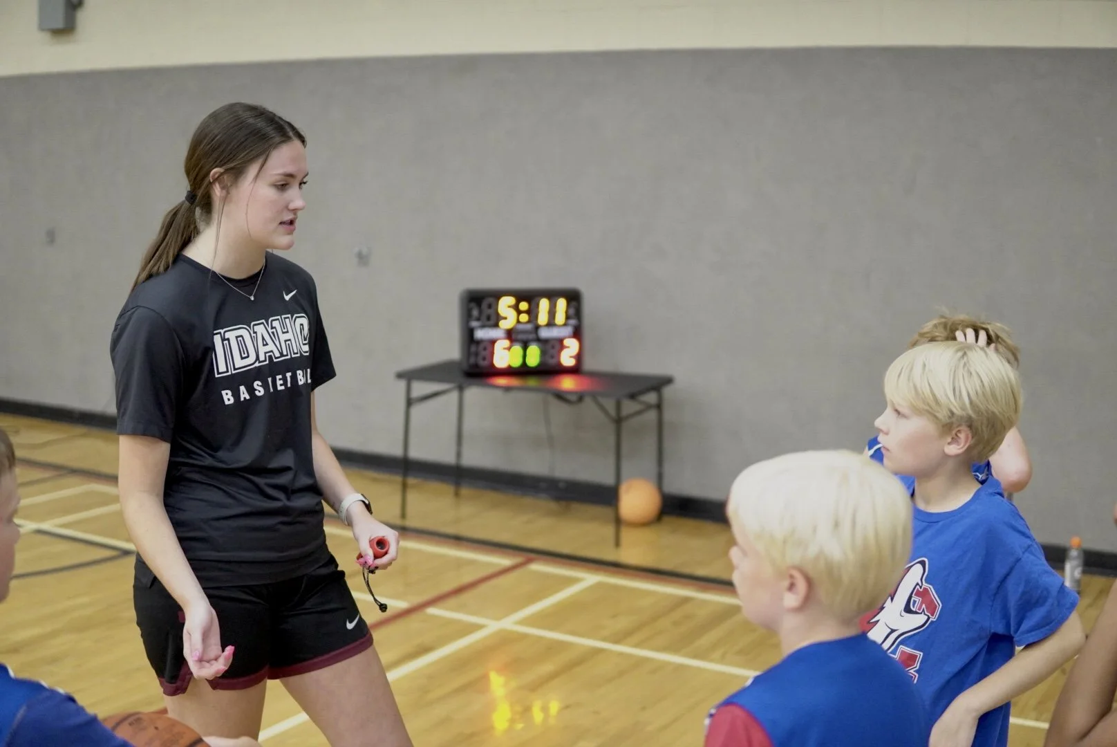 A young female basketball coach with long brown hair in a ponytail, wearing a black Idaho Basketball t-shirt and shorts, holding a stopwatch, speaking to a group of children in a gymnasium. There is a scoreboard in the background showing 5:11 minutes