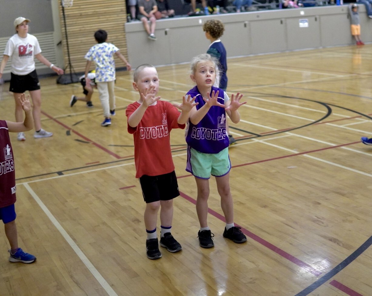 Children playing and participating in a gym class or sports activity indoors on a wooden basketball court.