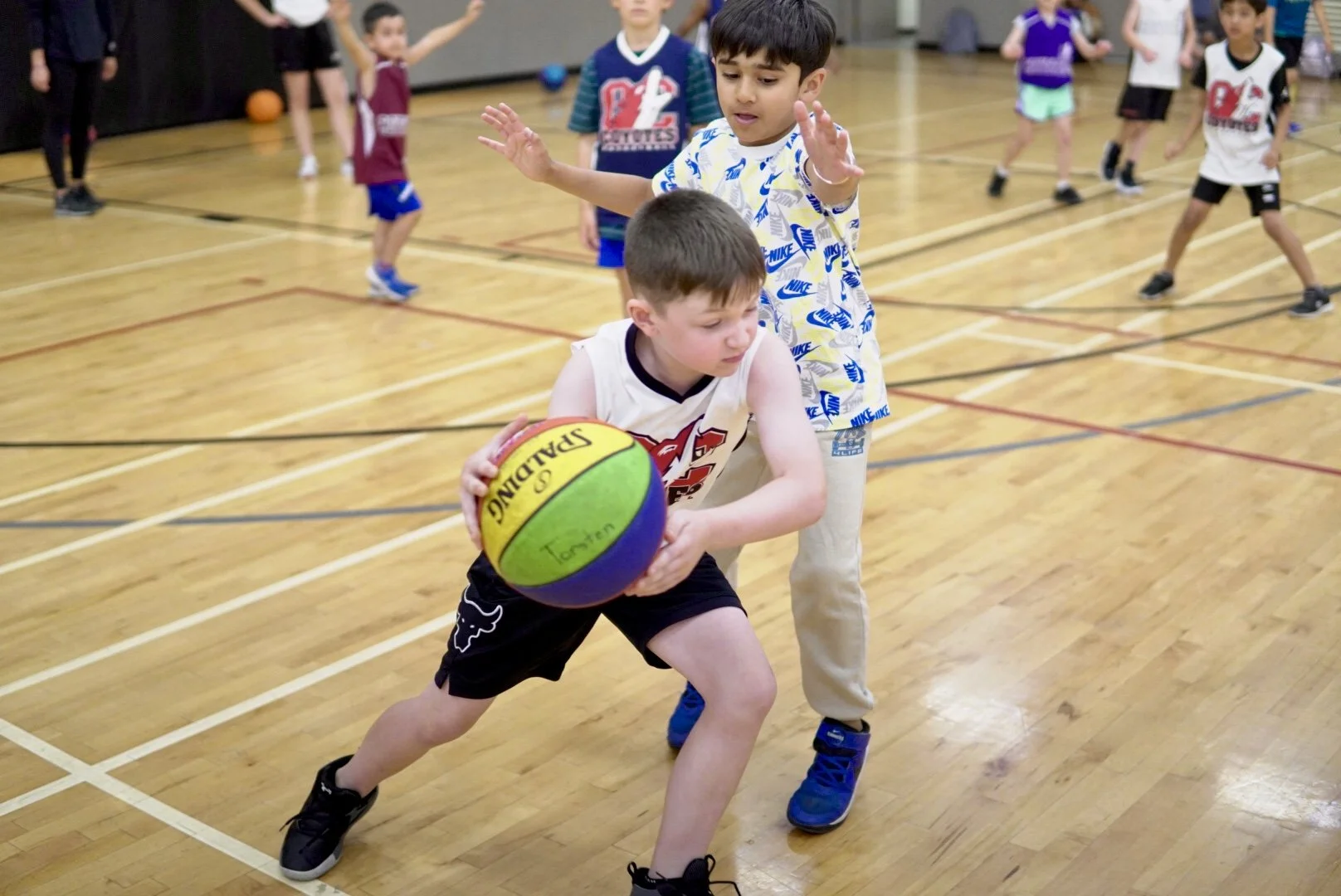 Two young boys playing basketball in a gym. The boy in front is holding a multicolored basketball, and the boy behind is defending. Several other children are in the background, participating in the game.