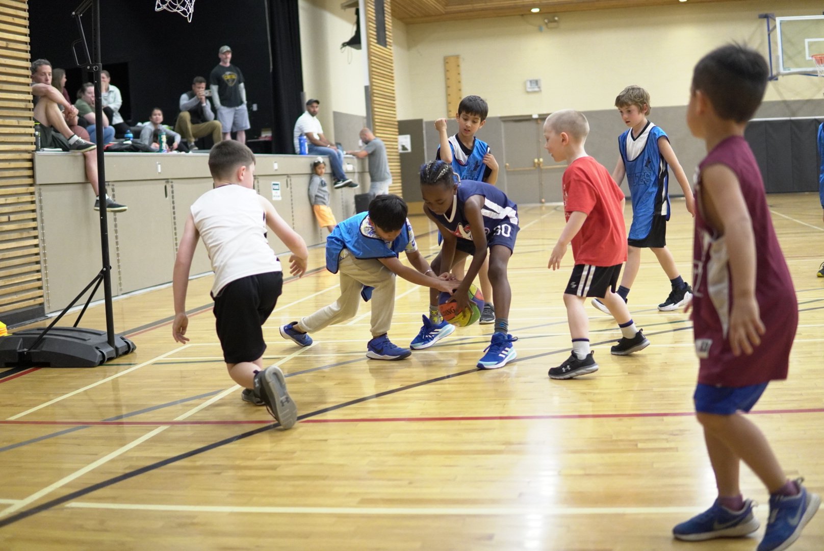 Kids playing basketball on an indoor court, with some children trying to grab the basketball while others are watching or moving around.