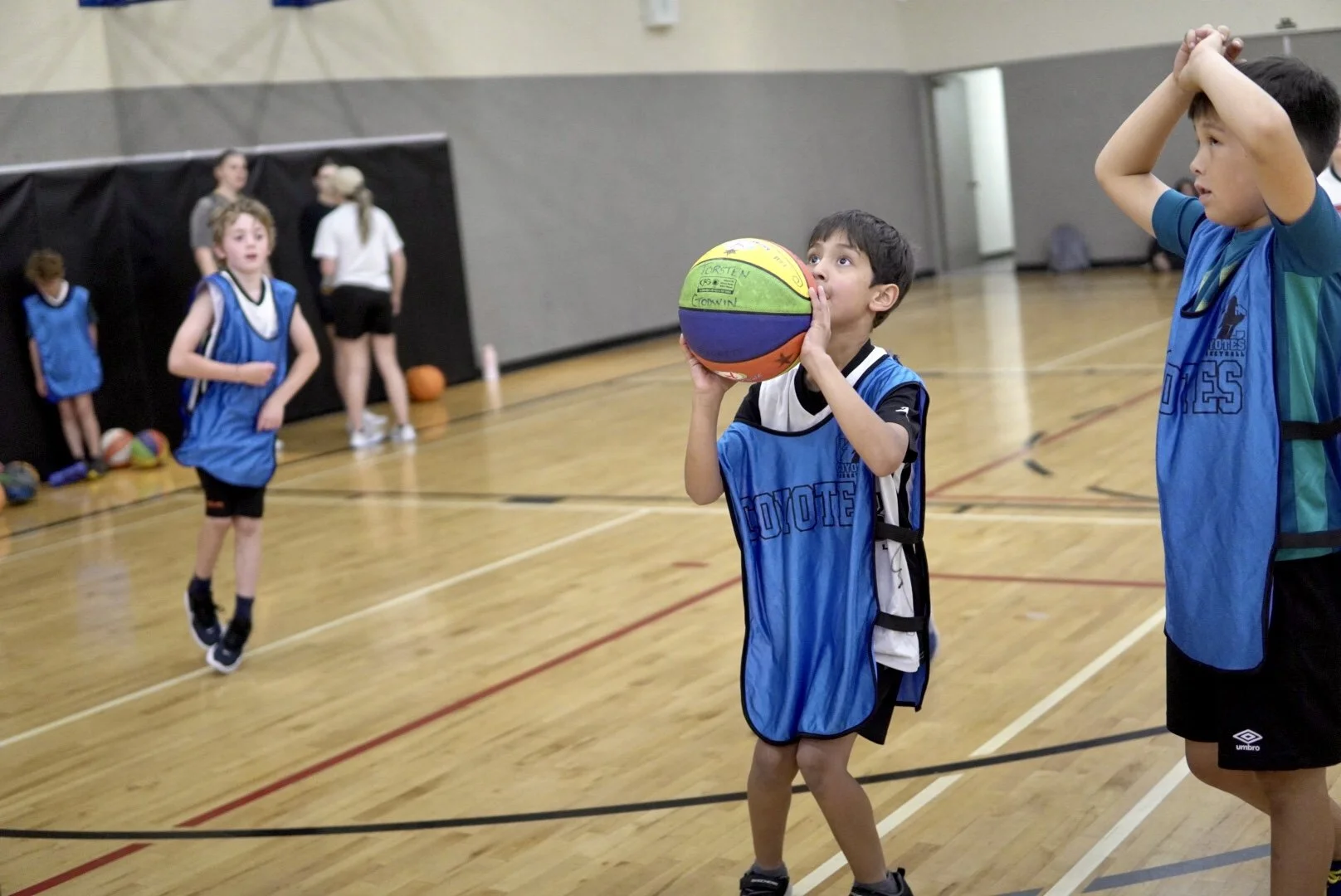 Young boys playing basketball in a gymnasium during practice or a game, with some kids in the background and one boy preparing to shoot the ball.